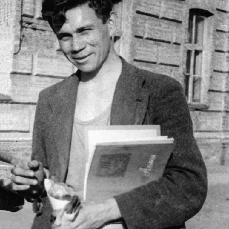 Photograph of a person standing outdoors in front of a brick building, holding books and a rolled-up paper. 