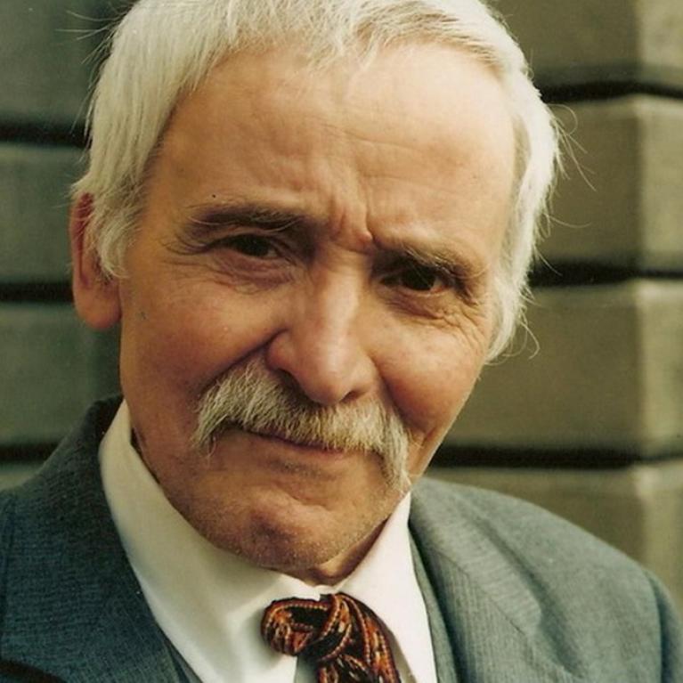 Photograph of an older man wearing a gray suit jacket, white shirt, and patterned tie standing in front of a stone building with horizontal lines. The setting suggests an outdoor urban environment with a formal or professional context.