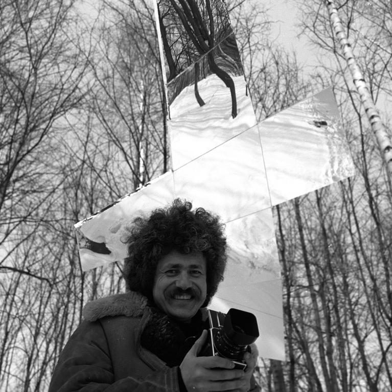 Photograph of a person holding a camera in a forest with leafless trees, standing in front of a large reflective cross structure. The cross reflects parts of the surrounding trees and sky, creating a striking contrast between natural elements and the shiny, angular surface.