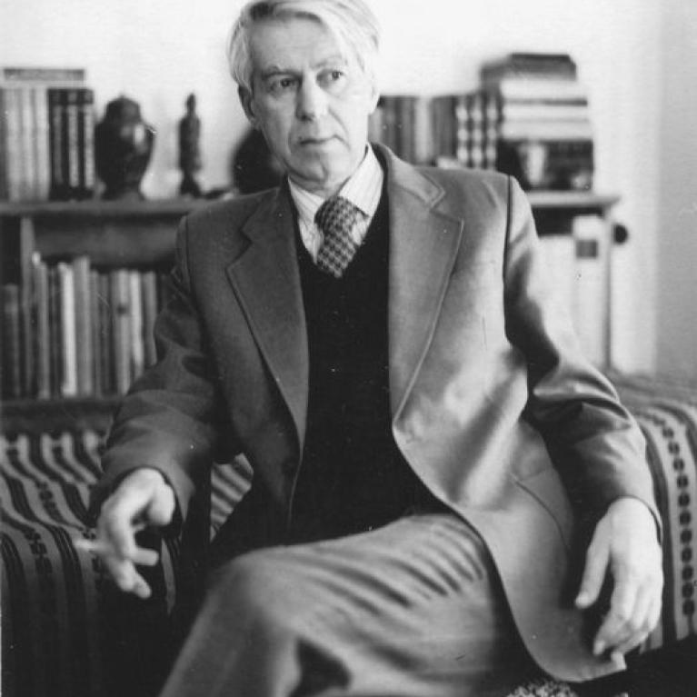 Black and white photograph of a man seated in a chair wearing a suit jacket, sweater, and tie, with a bookshelf filled with books and decorative items in the background. The setting suggests a formal or intellectual environment, highlighting a composed and relaxed posture.