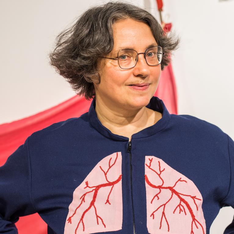 Photograph of a person wearing a navy blue jacket with a large pink lung design featuring red branching airways on the front. The background includes red and white fabric elements, suggesting a health or respiratory-related theme.