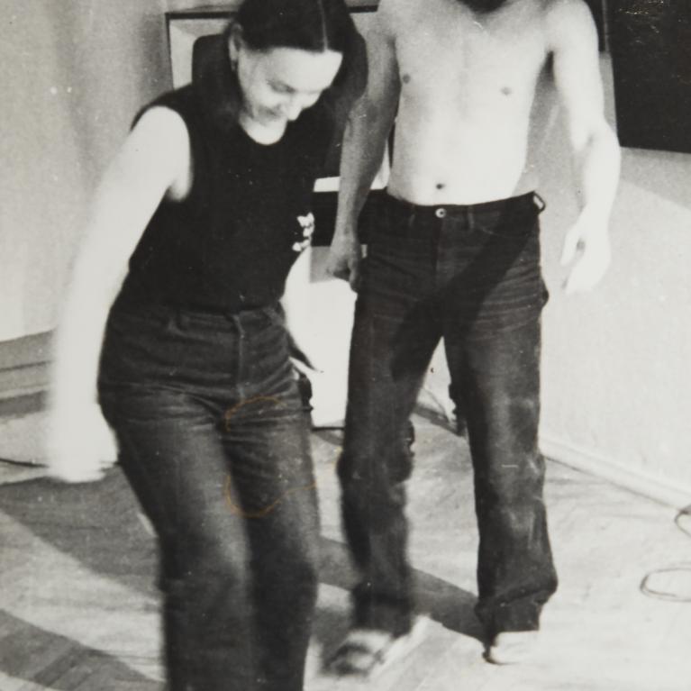 Black and white photograph showing a woman and a shirtless man standing indoors on a wooden floor, both wearing dark pants. The setting appears casual, with minimal background details including a curtain and a wall, suggesting a candid or informal moment.