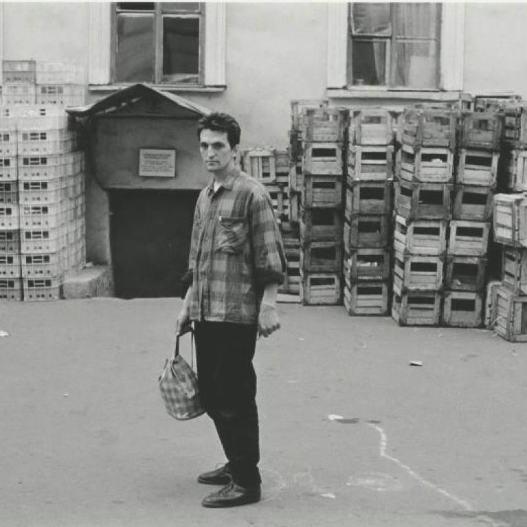 Photograph of a person standing outdoors in front of large stacks of empty crates arranged against a building wall. The person wears a plaid shirt and holds a bag, with windows visible on the building behind the crates.