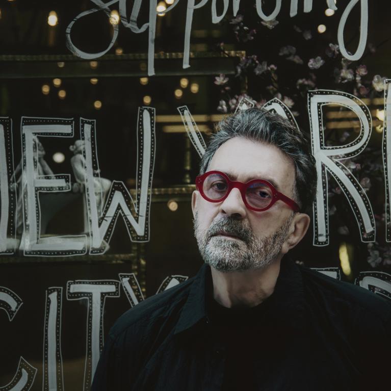 Photograph of a person standing in front of a glass window with large, handwritten-style text reading "NEW YORK CITY" and additional smaller text above and below. The scene is dimly lit with warm lights reflecting on the glass, creating an urban, nighttime atmosphere.