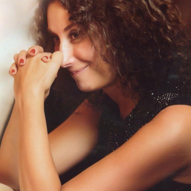 Photograph of a person with curly hair wearing a sleeveless black top, sitting at a table with hands clasped together in front of them. The setting appears to be indoors with warm lighting.