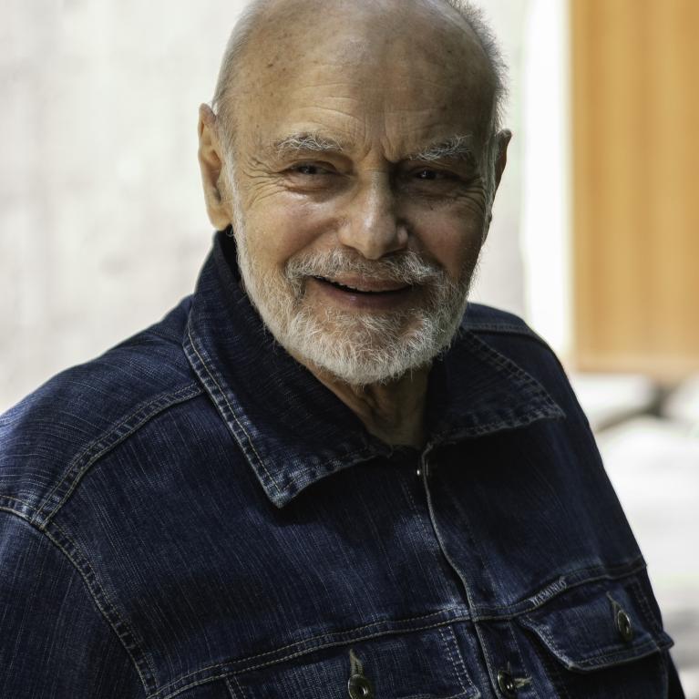Photograph of an elderly person wearing a dark blue denim jacket standing outdoors. Background includes a building with windows and an orange door, suggesting an urban or residential setting.