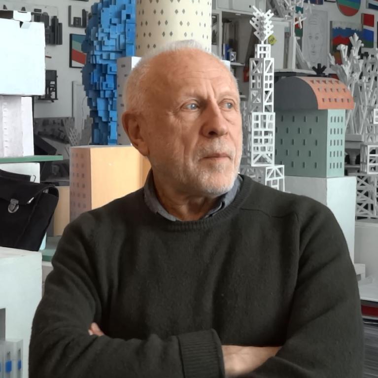 Photograph of a person sitting with arms crossed in front of architectural models and design prototypes. The background features various scale models in white, blue, and gray colors, showcasing different building structures and design elements.