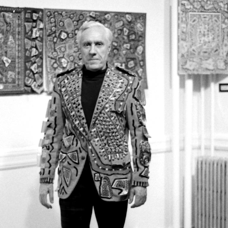Photograph of a person standing indoors wearing a uniquely patterned jacket featuring various geometric and abstract shapes. Background includes framed textile art pieces on walls and a radiator, suggesting an art gallery or exhibition setting.