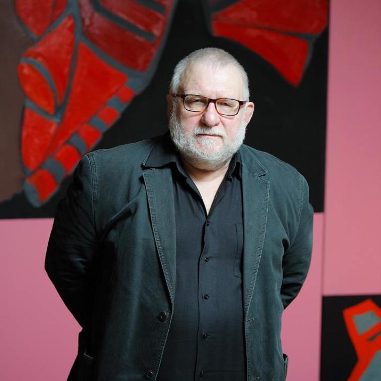 Photograph of a person standing indoors against a wall decorated with large, abstract red and black artwork. The person wears a dark blazer and shirt, positioned centrally with hands behind their back, highlighting a formal or professional setting.