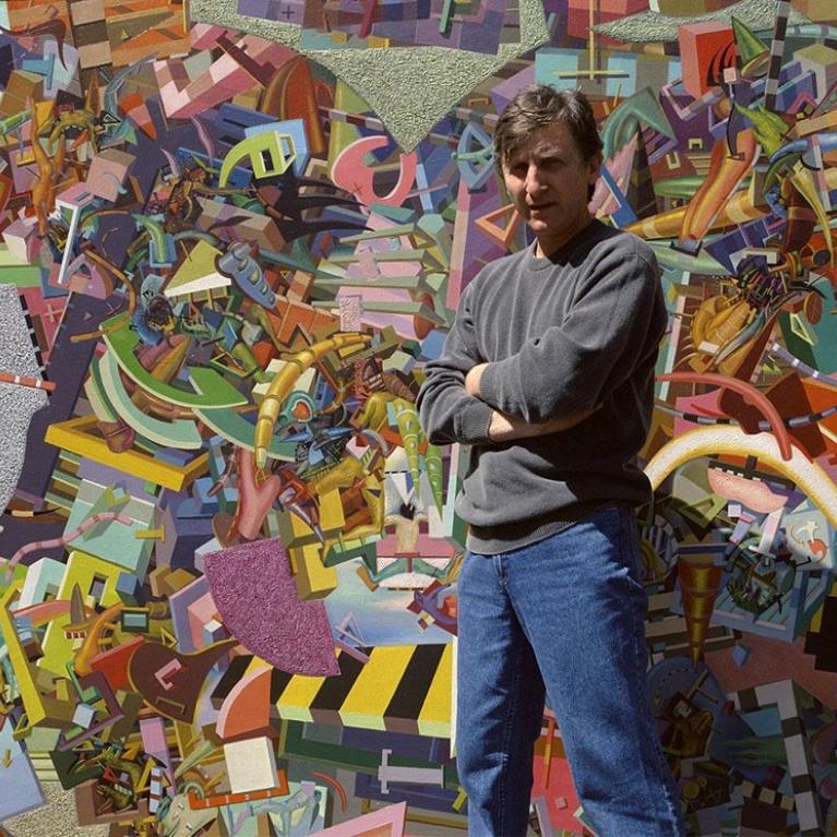 Photograph of a person standing in front of a large, colorful abstract mural composed of geometric shapes, curved lines, and varied textures in vibrant hues like purple, yellow, green, and pink. The mural features layered elements creating depth and complexity, with a mix of sharp angles and flowing forms that suggest dynamic movement and artistic expression.