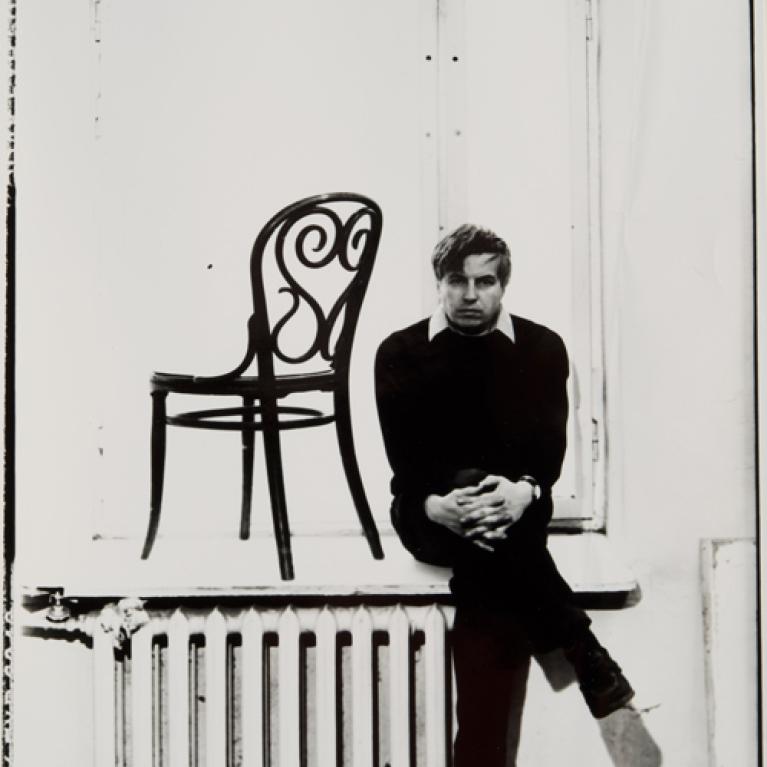Black and white photograph showing a person sitting on a windowsill next to a decorative chair placed on the sill. The composition highlights contrast between dark clothing and light background, emphasizing shapes and shadows around the radiator and window frame.