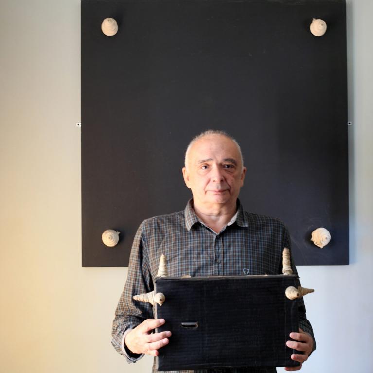 Photograph of a person standing in front of a large black square panel mounted on a wall, holding a smaller black rectangular object with four protruding spikes. The panel and object contrast with the light-colored wall, and the spikes on both items create a visual connection between them.