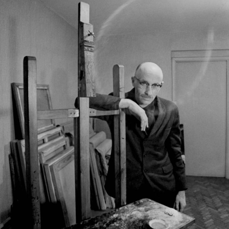 Black and white photograph of a person standing in an art studio, leaning on an easel surrounded by stacked canvases and a paint-splattered table. The setting features a bare light bulb hanging from the ceiling and a closed door in the background, emphasizing a minimalist and focused creative environment.