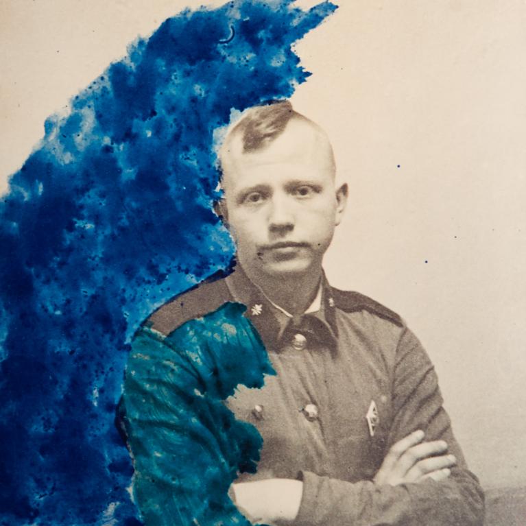 A photograph of a person in a military-style uniform with arms crossed, partially obscured by an abstract blue and red paint overlay. The paint creates a dynamic contrast against the grayscale figure, blending artistic expression with historical portraiture.