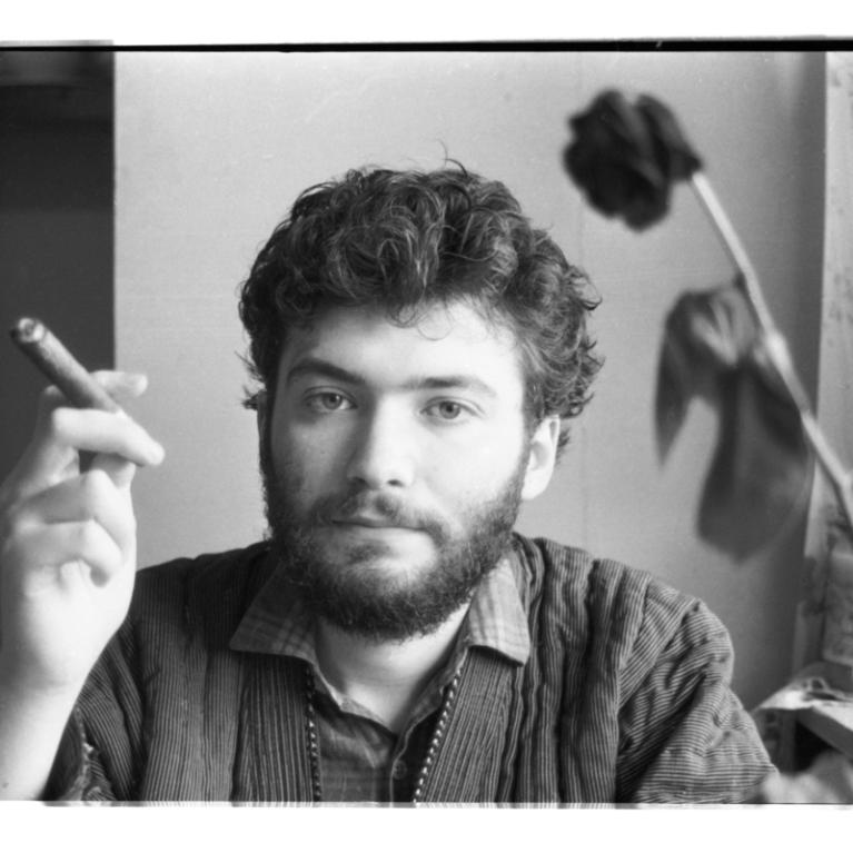 Black and white photograph of a person sitting indoors, holding a cigarette in their right hand. A wilted flower is visible in the foreground on the right side, with a curtain and tiled wall in the background.