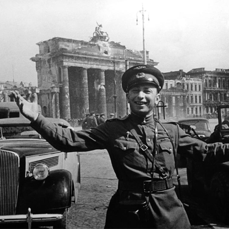Black and white photograph showing a uniformed soldier standing with arms outstretched in front of vintage cars and a large classical building with columns. The scene captures a historical urban setting, likely the end of WWII, with architectural details and period vehicles emphasizing the era.