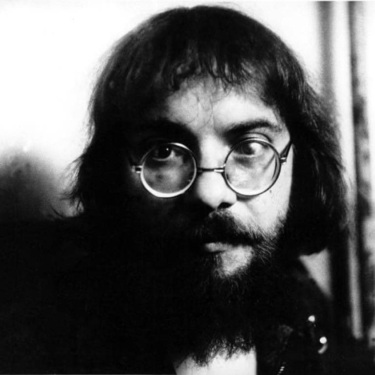 Photograph of a person with long, dark hair and a beard, wearing round glasses and a dark jacket, set against a light background. The image is in black and white, highlighting contrast between the subject and background.