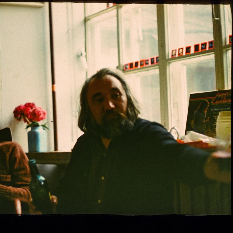 Photograph of a person sitting indoors near a window with a vase of red flowers on a table to their left. The person is holding a bottle and appears to be gesturing with their other arm, with a collection of small framed pictures or cards displayed along the window sill.