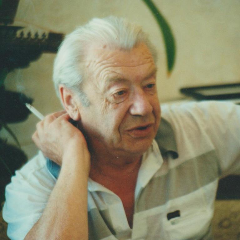 Photograph of an older man with white hair wearing a light-colored striped polo shirt, seated indoors with a cigarette in his right hand. Background includes a dark piece of furniture and some green plant leaves.