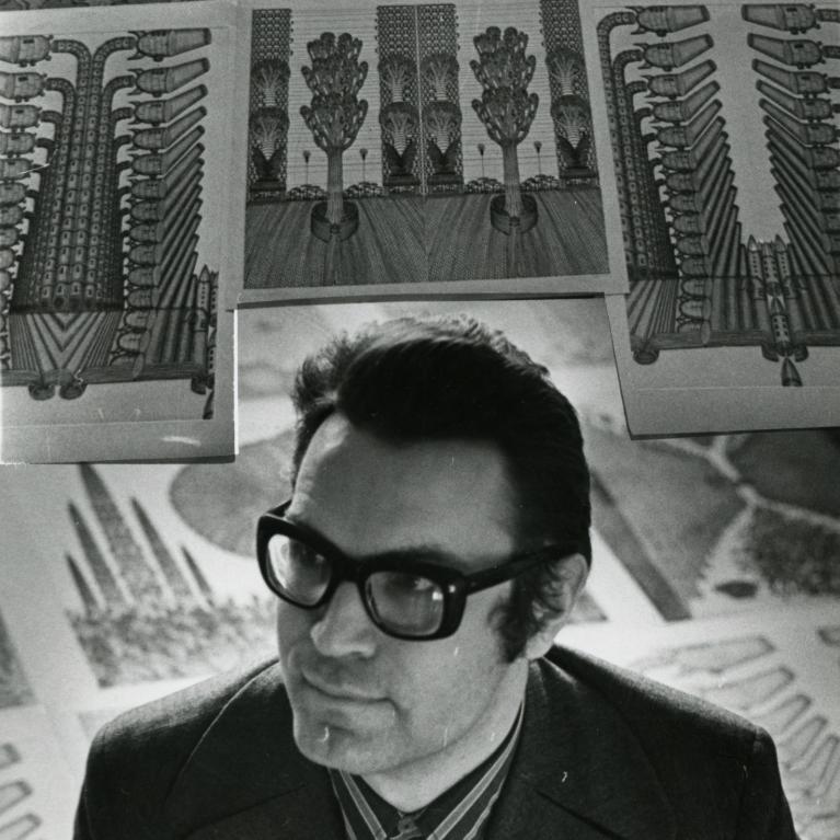 Black and white photograph of a person seated in front of a wall covered with detailed botanical or scientific illustrations featuring symmetrical plant or insect-like structures with intricate patterns. Illustrations include vertical and horizontal elements with repetitive shapes resembling leaves, seeds, or mechanical parts, suggesting a study or display of natural forms or designs.