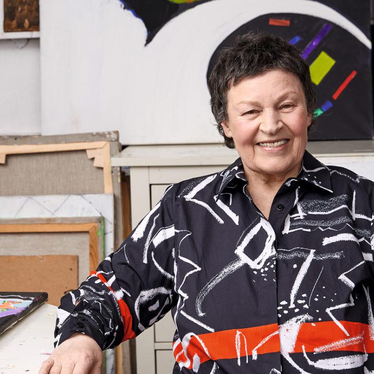 Photograph of a person seated in an art studio surrounded by colorful abstract paintings with geometric shapes. The person wears a black shirt with white and red abstract patterns, blending with the vibrant artwork in the background.