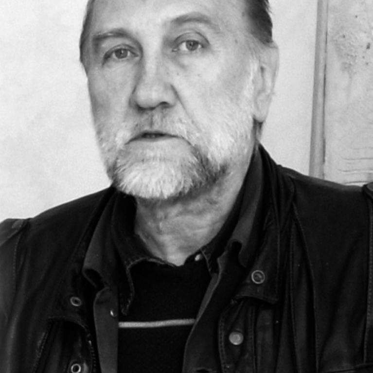 Black and white photograph of an older man wearing a leather jacket over a striped sweater, standing against a plain wall. The photo captures a casual, candid moment with a focus on clothing texture and contrast.
