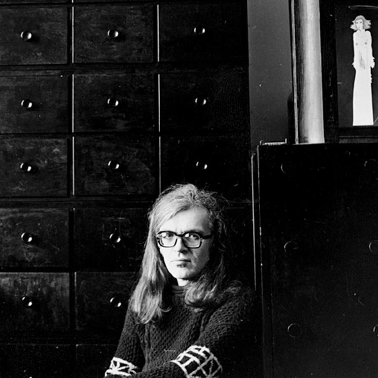 Black and white photograph showing a person with long hair sitting in front of a large wooden cabinet with multiple drawers