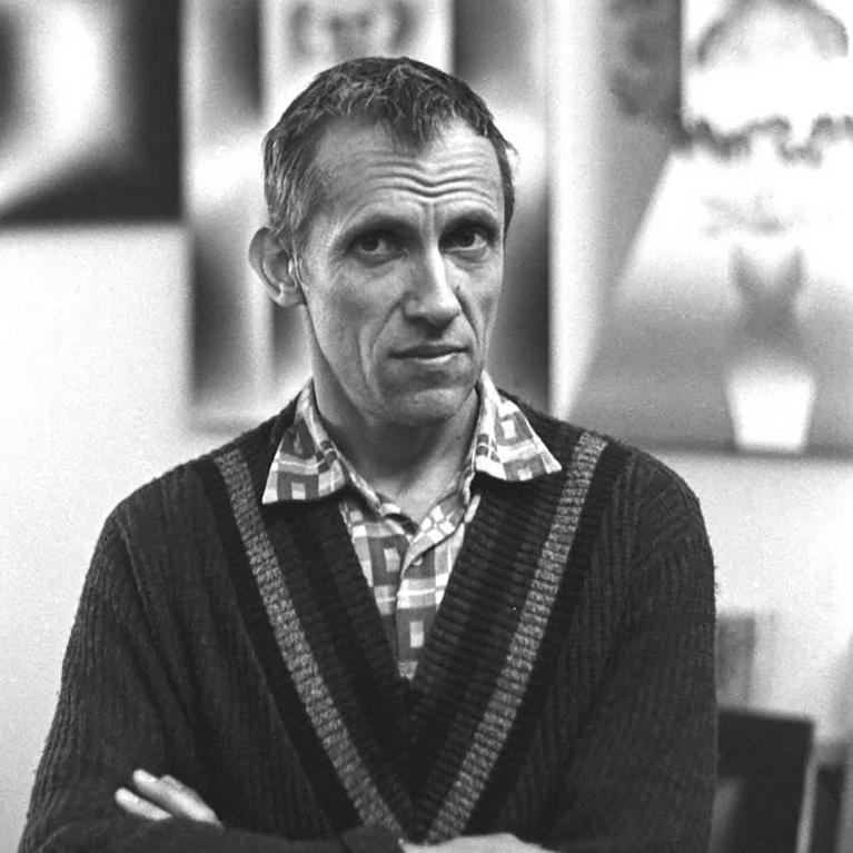 Black and white photograph of a man standing indoors with arms crossed, wearing a patterned shirt under a dark cardigan. Background features blurred abstract artwork and framed pictures on a wall.