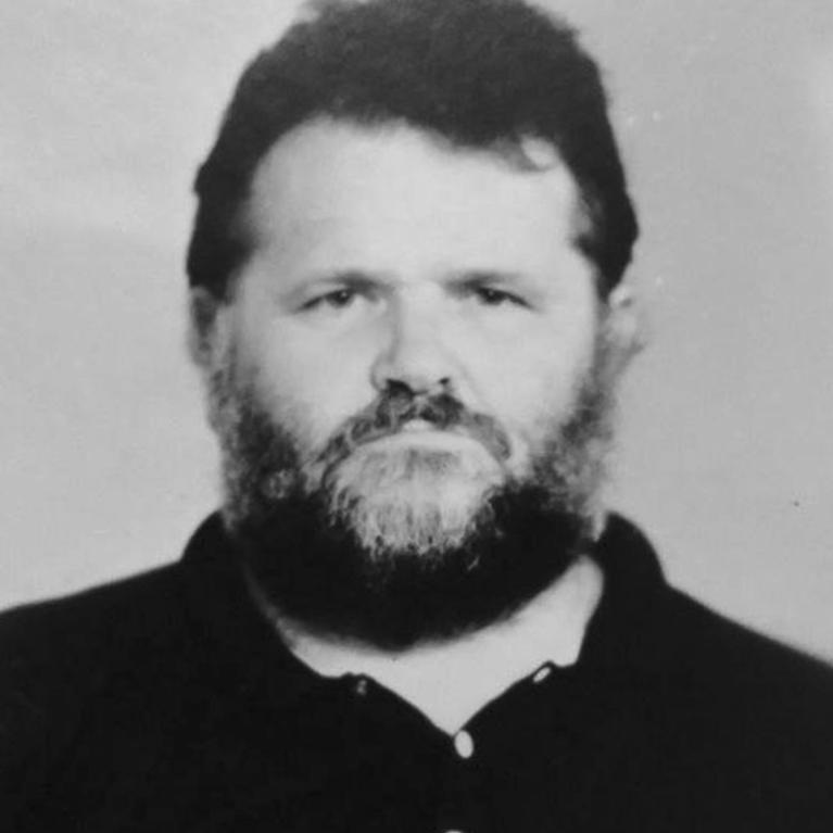 Photograph of a person with bearded face wearing a dark collared shirt with two visible buttons. The grayscale portrait focuses on the upper torso and head against a plain background.
