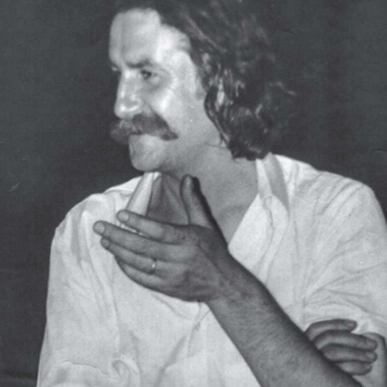 Black and white photograph of a person with moustache wearing a short-sleeve button-up shirt with arms crossed and one hand raised near the face. 