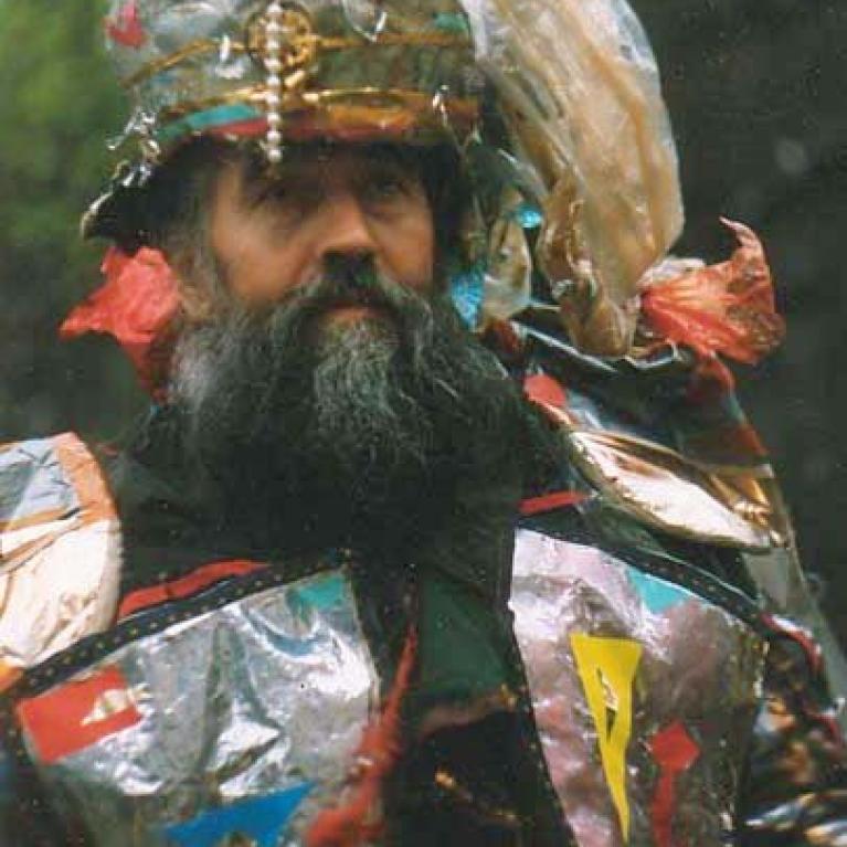 Photograph of a person wearing a colorful, reflective costume made from metallic and plastic materials, featuring various shapes and colors like red, blue, yellow, and silver. The outfit includes a headpiece with layered elements and a beard visible beneath, set against an outdoor background.