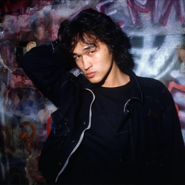 Photograph of a person with dark curly hair wearing a black jacket and shirt, posing with one arm raised behind their head against a colorful graffiti-covered wall. 