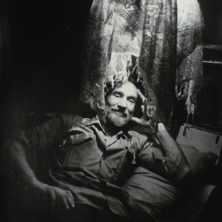 A black and white photograph showing a person sitting in a chair with a patterned cloth draped over the hanging lamp. The setting appears dimly lit, creating a mysterious and eerie atmosphere with strong contrasts between light and shadow.