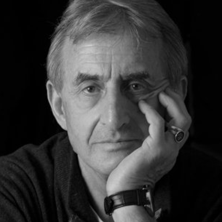 A black and white photograph showing a person with light-colored hair resting their head on their hand, wearing a dark zip-up sweater and a wristwatch. The background is dark, emphasizing the subject's contemplative pose and facial features.