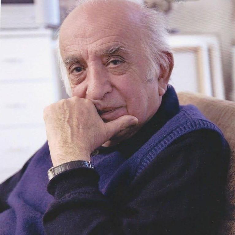 Photograph of an elderly person sitting on a couch wearing a dark blue sweater and a wristwatch, with their hand resting near their face. Background shows a blurred indoor setting with stacked canvases on stretchers.