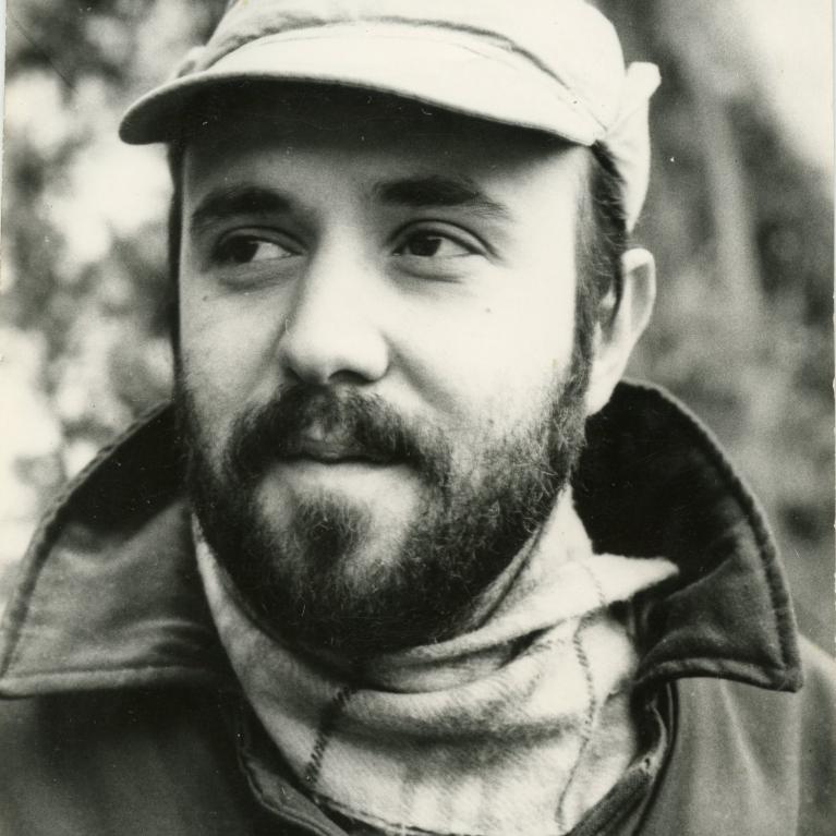 Photograph of a person wearing a cap, scarf, and coat, standing outdoors with blurred trees in background. The photo is in black and white