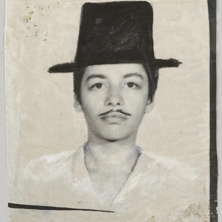 A black and white photograph of a person with scribbled moustache and a tall, wide-brimmed hat. The photo has a vintage, worn appearance with visible creases and discoloration around edges.