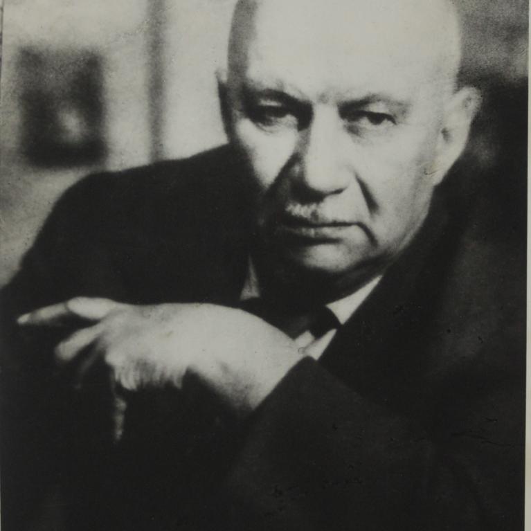 Black and white photograph showing a person in a suit. The person is positioned with one arm bent and hand near their face, set against a blurred background.