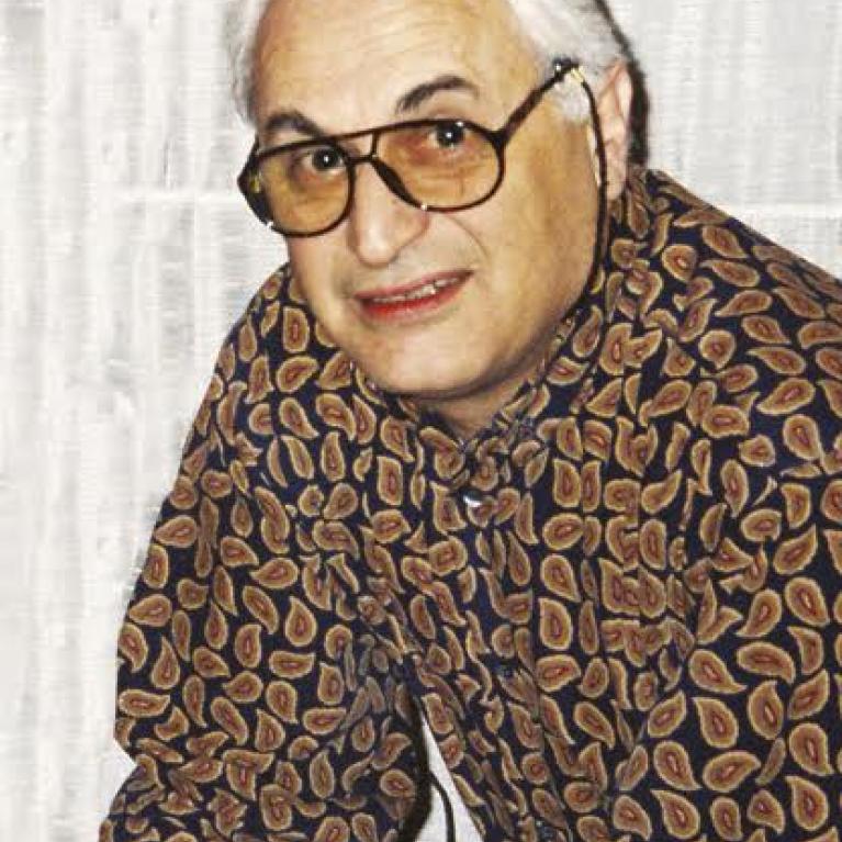 Photograph of a person sitting against a light-colored curtain background. The person wears a patterned long-sleeve shirt with a leaf-like design in brown and orange tones.