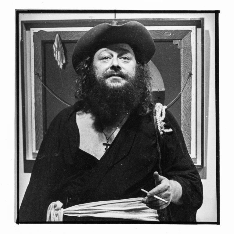 Black and white photograph of a person dressed in dark, loose clothing with a beret, holding a cigarette in one hand and a cloth in the other, standing in front of a framed artwork. The person has a long beard and wears a cross necklace, with a rope draped over one shoulder, creating a dramatic and artistic portrait.