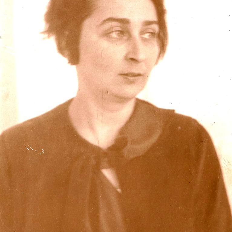 Photograph of a person wearing a dark-colored garment with a bow detail at the neckline, set against a plain light background. The photo has a sepia tone, giving it a vintage appearance.
