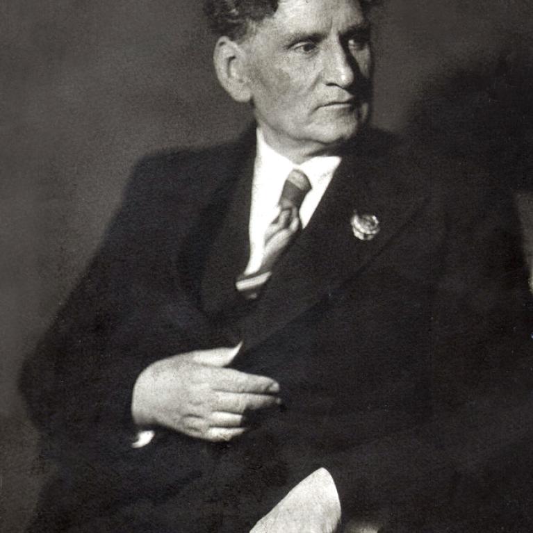 Photograph of a seated person wearing a dark suit, white shirt, and striped tie, with a decorative pin on the lapel. The person has curly hair and is posed with one hand resting on their chest and the other on a chair arm.