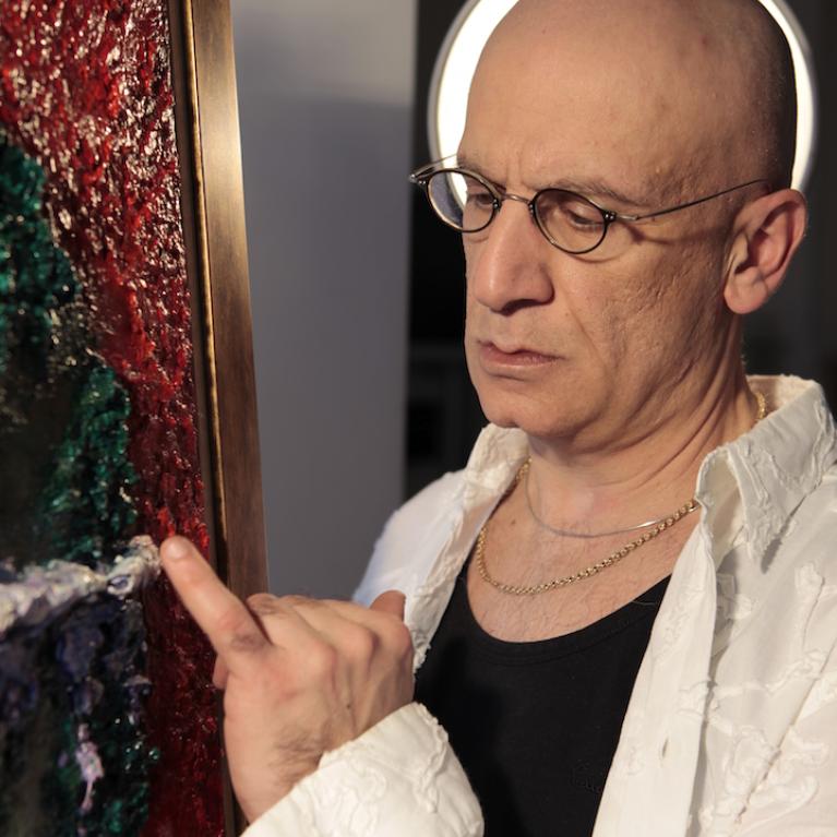 Photograph of a person with a shaved head and glasses pointing at a textured, colorful painting with red, green, and purple hues in a gold frame. Person wears a white patterned jacket over a black shirt and gold chains, with a circular light source behind their head creating a halo effect.
