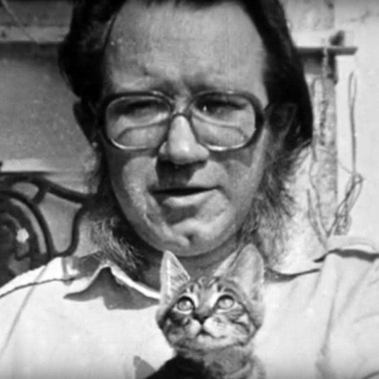 Black and white photograph showing a person holding a small tabby kitten looking upward. Background includes a metal railing and some indistinct objects, suggesting an outdoor or porch setting.