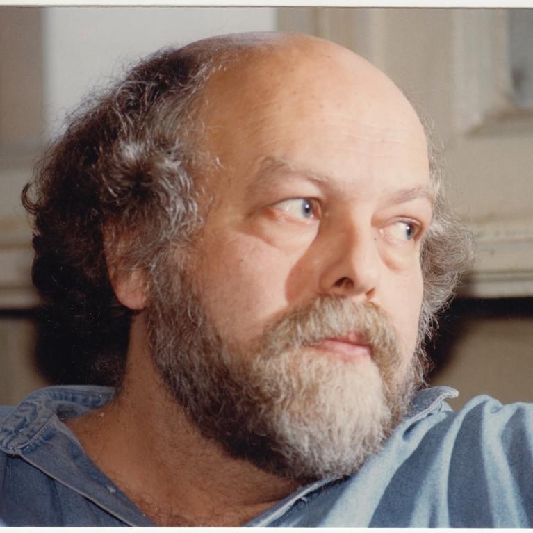 Photograph of a man with curly hair and a beard wearing a blue shirt, sitting indoors near a window with white curtains. The setting appears casual and relaxed, with neutral tones in the background.