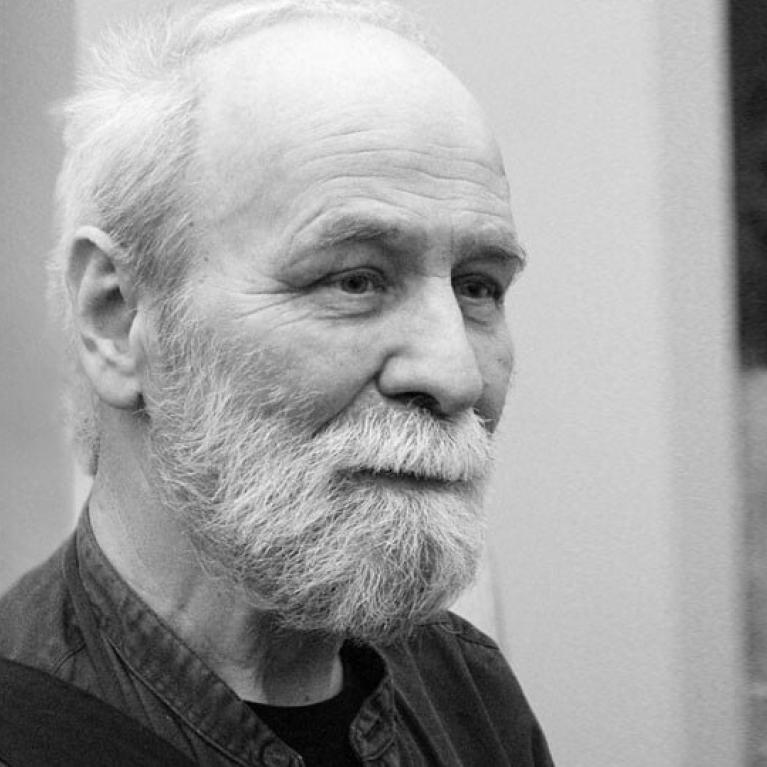 Black and white photograph showing an elderly person with white hair wearing a dark shirt. The background includes a blurred wall and a partially visible framed artwork.