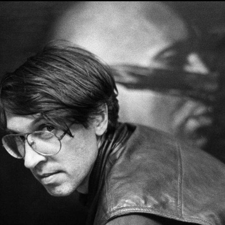 Black and white photograph showing a person with short hair wearing a leather jacket, facing sideways. Background features a large, partially obscured portrait.