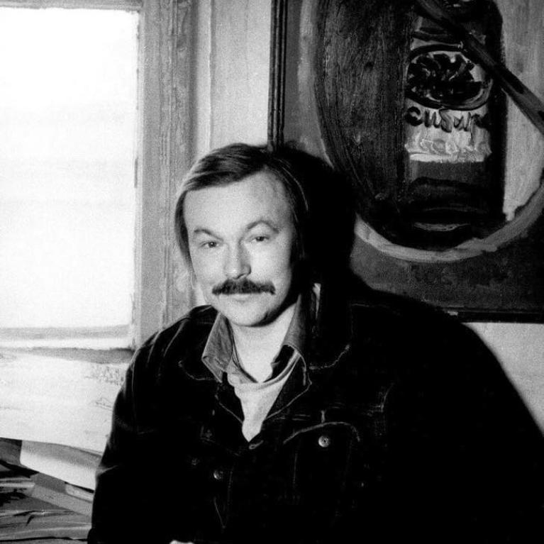 A black and white photograph showing a person sitting indoors near a window with light streaming in. Behind the person is a framed abstract painting featuring dark and light shapes, adding artistic context to the setting.