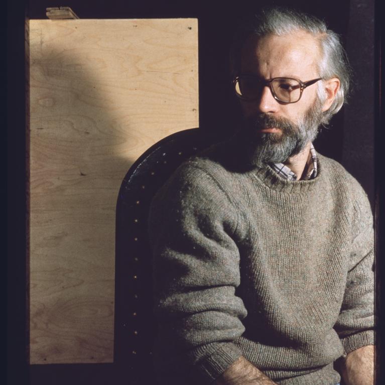 Photograph of a person sitting on a chair wearing a gray sweater and blue jeans. The background features a wooden panel partially illuminated by light, creating contrast with the dark surroundings.