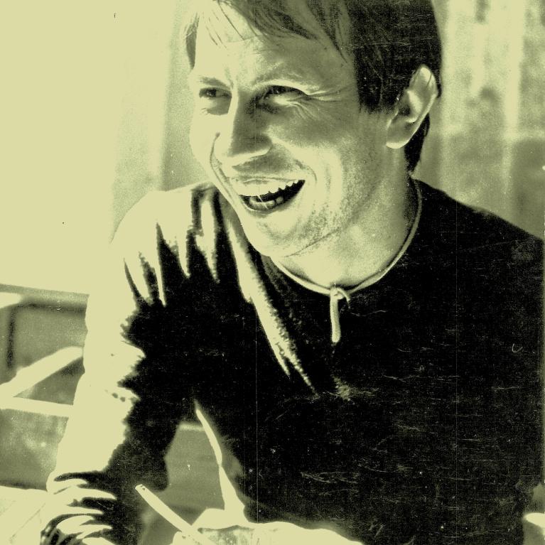 Photograph of a person sitting at a table, his smiling face highlighted by sun light coming from the window. The image has a vintage, grainy texture and muted color tones, suggesting an old photo.